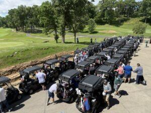 Row of black golf carts parked side by side on a paved path at a golf course, with players and bags nearby.