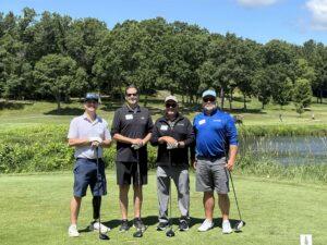 Four golfers stand together on a sunny course, posing with their clubs in front of a green, tree-filled backdrop.
