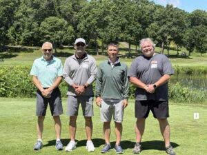Four men in golf attire pose together on a sunny golf course with trees in the background and name tags on their shirts.