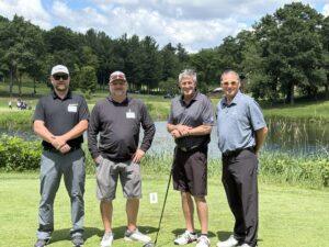 Four men stand together on a golf course posing for a photo, holding golf clubs with a pond and trees in the background.