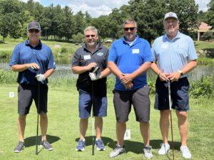 Four men posing with golf clubs on a sunny golf course, wearing polo shirts and caps.