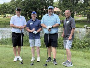 Four men in golf attire posing with clubs on a green near a pond at a sunny golf course.