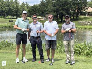 Four men stand together on a golf course near a pond, each holding a golf club and smiling at the camera.