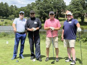 Four men posing on a sunny golf course with clubs, standing in a row by a pond.