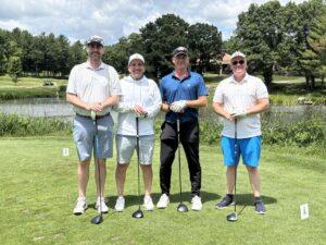 Four men in golf attire posing with clubs on a green golf course, with trees and a pond in the background.