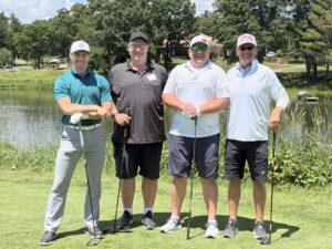 Four men posing with golf clubs on a sunny golf course by a pond.