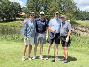 Four men in golf attire pose together on a sunny golf course with a pond and green landscaping in the background.