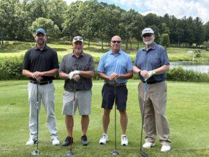 Four men standing on a golf course holding clubs, posing for a photo with trees and a pond in the background on a sunny day.