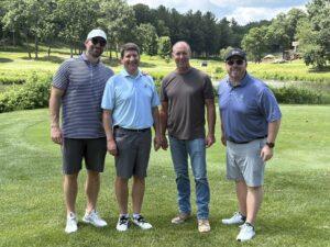 Four men standing side by side on a sunny golf course, posing near a pond in the background.