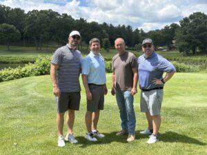 Four men stand together on a sunny golf course, smiling, with trees and a water hazard in the background.