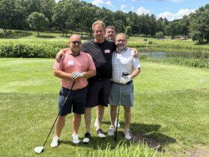 Four men stand together on a sunny golf course, holding their clubs and smiling, with a pond and trees in the background.