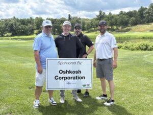Four men on a golf course posing with a sponsor sign reading Oshkosh Corporation