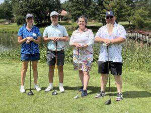 Four golfers stand side by side on a sunny golf course, their clubs resting on the ground as they pose for the photo.