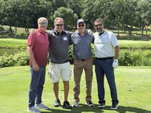 Four men in golf attire pose with arms around each other's shoulders on a sunny course.