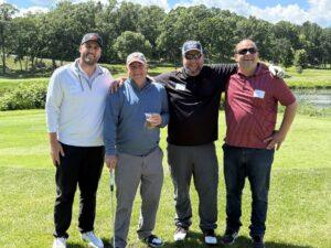 Four men posing together on a sunny golf course, smiling with green fairways and trees in the background.
