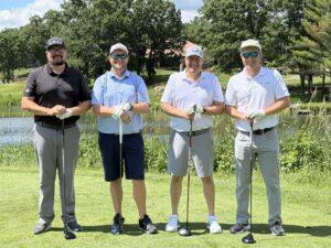 Four male golfers stand side by side on a sunny golf course, each holding a club by a small pond behind them.