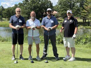 Four men stand with golf clubs on a lush course posing for a group photo by a pond.