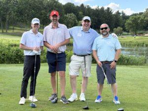 Four men posing with golf clubs on a greenspace, trees and a pond in the background during a sunny day.