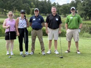 Five adults pose on a golf course, each holding a club and smiling, with trees and a pond in the background.