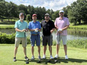 Four men in golf attire posing with clubs on a sunny golf course by a pond and trees behind them.