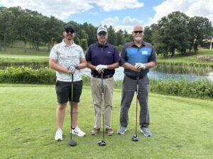 Three men in golf attire posing with clubs on a sunny golf course, a pond and trees behind them.