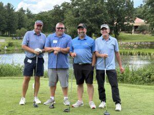 Four men in polo shirts and caps posing with golf clubs on a green course near a pond on a sunny day.