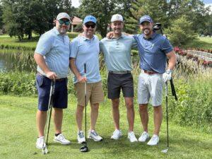 Four friends in golf attire pose with arms around each other on a sunny golf course, holding their clubs near a pond.