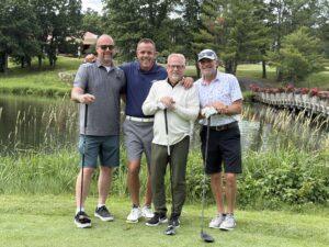 Four men stand together on a golf course, each holding a club, with a pond, a wooden bridge, and blooming flowers in the background.