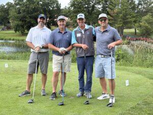 Four men with golf clubs posing for a group photo on a sunny golf course with trees in the background and a small pond nearby.