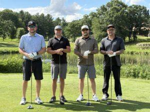 Four men stand side by side on a golf course, posing with their clubs under a sunny blue sky.