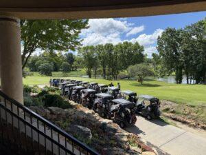 Line of golf carts parked along a sidewalk at a golf course, with players loading gear near a landscaped entrance and a lake in the background.