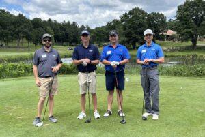 Four men pose with golf clubs on a manicured course, under a partly cloudy sky and trees in the background.