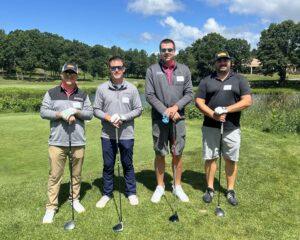 Four men in golf attire posing with clubs on a sunny, green golf course with trees in the background.