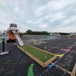Temporary putting-green setup in a parking lot for an outdoor event, with cones, tape, and a large orange-and-white maintenance vehicle nearby.