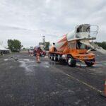 Orange concrete mixer truck with chute extended at a work zone; tents line the left, orange cones mark the area under an overcast sky.