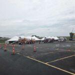 Row of white tents in a parking lot with a large orange rocket-like aircraft on a trailer and orange traffic cones marking lanes
