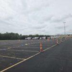 Wide empty parking lot lined with orange traffic cones and a row of cement mixer trucks in the distance under a cloudy sky.