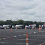 Row of cement mixer trucks parked in a lot with orange traffic cones and colorful ribbons in the foreground under a cloudy sky.