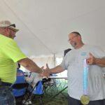 Two men shake hands in a tent at an outdoor award event; one wears a neon safety shirt and cap while the other wears a gray T-shirt and a gold medal on a ribbon.