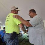 Two men hug under a white tent at an outdoor event, one in a neon shirt and jeans, the other in a gray T-shirt, with medals and trophies nearby.