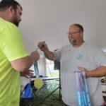Man in a gray t-shirt hands a medal to a man in a neon yellow shirt at an outdoor awards tent, ribbons and chairs visible in the background.