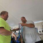 Man in a neon shirt hands a medal to a man in a gray T‑shirt under a tent; trophies visible on a table behind them.