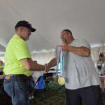 Two men shake hands under a tent; one in a neon shirt hands over medals and trophies to the other man smiling nearby.
