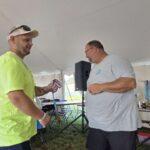 Two men under a tent exchange a medal during an outdoor event, neon-shirted volunteer handing a ribboned award to a balding man with glasses.