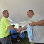 Two men shake hands in a tent at an awards event; one wears a neon lime shirt, the other a gray T-shirt with a ribbon and medal hanging from his neck.