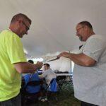 Two men under a white tent exchange a small item while others sit at tables in the background.