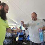 Bearded man in a neon shirt hands a blue ribbon medal to another man in a gray shirt under a white tent.