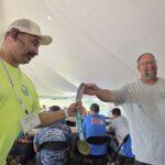 Two men under a tent exchange a ribbon or award; man in lime shirt hands a medal to man in gray shirt, smiling event scene in background.