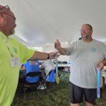 Man in neon yellow shirt hands a medal on a ribbon to a smiling man in a gray shirt under a tent; trophy table and spectators in the background.