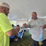 Two men under a white event tent exchange medals; one wears a neon yellow shirt, the other a gray t‑shirt with ribbons and medals hanging from his neck.